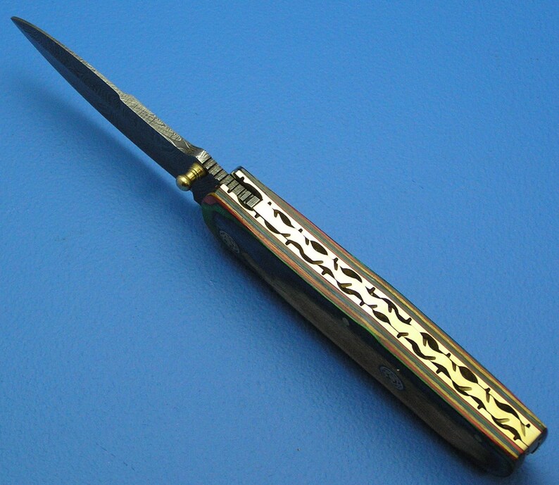May include: A handcrafted folding knife with a dark blade and a handle featuring a gold-colored, leaf-like design. The handle has a dark wood appearance with gold accents. The knife is set against a blue background.