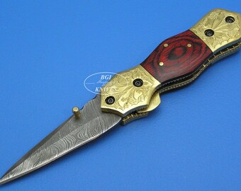 May include: A gold and red folding knife with a patterned blade and a wood handle. The knife has a decorative design on the handle and a small pin on the side.