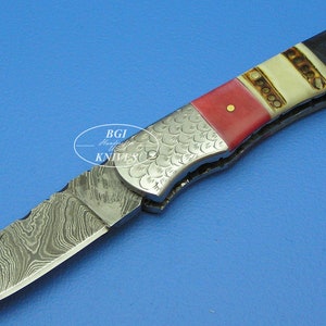 Puede incluir: Un cuchillo plegable de bolsillo con una hoja de acero Damasco y un mango rojo, blanco y azul. El mango está hecho de hueso y tiene un patrón decorativo.