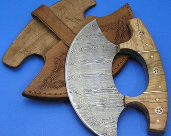 Op de afbeelding: Een gebogen, zilverkleurig mes met een houten handvat. Het handvat heeft een ring voor de duim en twee rode edelstenen. Het mes zit in een bruine leren schede.