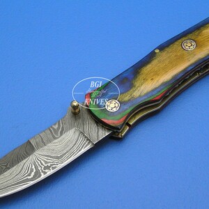 May include: A folding knife with a patterned blade and a handle featuring a colorful wood grain design. The handle has gold-colored accents and small decorative rivets. The knife is set against a blue background.