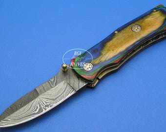 May include: A folding knife with a patterned blade and a handle featuring a colorful wood grain design. The handle has gold-colored accents and small decorative rivets. The knife is set against a blue background.