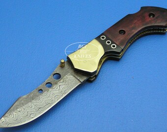 May include: A folding knife with a patterned steel blade and a wooden handle. The handle is a dark red color with a brass accent. The blade has a decorative pattern and a small hole near the tip.