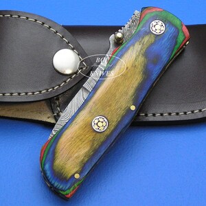 May include: A folding knife with a colorful wooden handle in shades of blue, yellow, and green, accented with brass rivets and a patterned blade. The knife rests on a brown leather sheath with a snap closure.