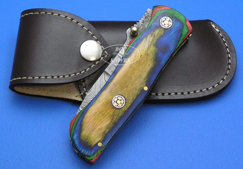 May include: A folding knife with a colorful wooden handle in shades of blue, yellow, and green, accented with brass rivets and a patterned blade. The knife rests on a brown leather sheath with a snap closure.