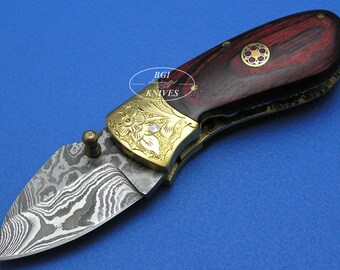 May include: A small pocket knife with a patterned steel blade and a red wood handle. The handle has a gold inlay with a floral design and a small diamond-shaped stone.