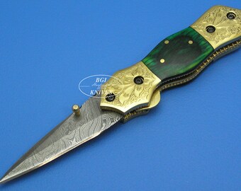 May include: A gold and green folding knife with a patterned blade and a wood handle. The knife is closed and has a decorative design on the handle.