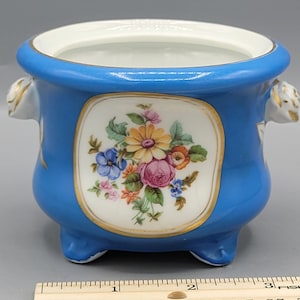 Könnte beinhalten: Ein kleiner blauer Porzellan-Pflanztopf mit einem weißen Blumenmuster und goldenen Akzenten. Der Topf hat zwei Griffe und drei Füße.