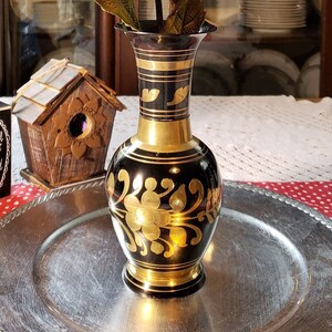 May include: A black and gold vase with floral designs sits on a silver tray. The vase is filled with a bouquet of orange silk flowers.