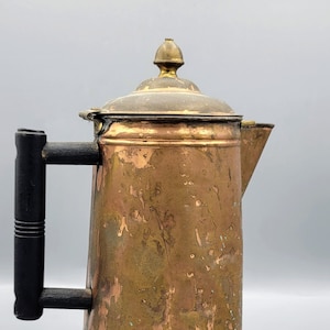 Puede incluir: Una cafetera de cobre vintage con un mango de madera y una tapa de latón. La cafetera tiene un aspecto desgastado y envejecido con un acabado de pátina.