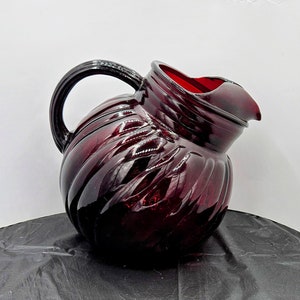 Puede incluir: Una jarra de vidrio de color rojo oscuro con un diseño acanalado. La jarra tiene un cuerpo redondeado y un asa.