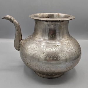 Puede incluir: Una tetera de metal de color plateado con un diseño grabado ornamentado. La tetera tiene un cuerpo redondeado y un pico con un asa curva.