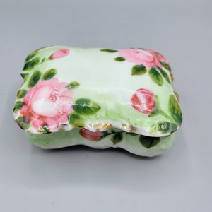 May include: A small, white porcelain trinket box with a green and pink floral design. The box has a slightly rounded, pillow-like shape.