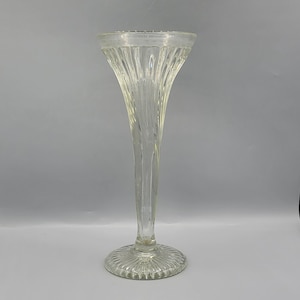 Puede incluir: Un jarrón de vidrio transparente con un tallo alto y delgado y una boca ancha y acampanada. El jarrón tiene un diseño texturizado y acanalado.