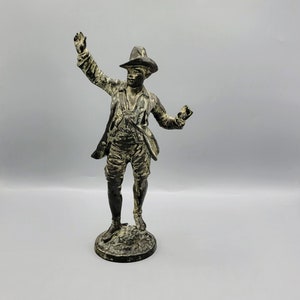 May include: A bronze figurine of a man wearing a hat and a long coat. The figurine is standing on a small base.