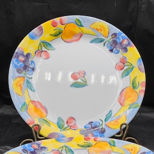 May include: A set of six white ceramic dinner plates with a colourful fruit design. The plates feature a yellow background with blue, red and orange fruits and leaves.