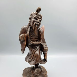 May include: A dark brown wooden sculpture of a man with a long beard and a long robe. The sculpture is standing on a wooden base.