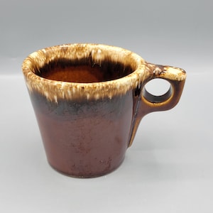 Può includere: Una tazza in ceramica smaltata marrone e bianca con un manico arrotondato. La tazza ha una smaltatura marrone con striature bianche e un bordo bianco.