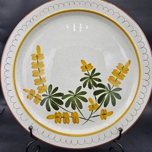 May include: A white ceramic dinner plate with a brown rim and a scalloped edge. The plate features a hand-painted design of yellow and green flowers.