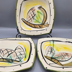 Può includere: Tre piatti quadrati in ceramica con fondo bianco e bordo verde. Ogni piatto presenta un design astratto stilizzato in tonalità di marrone, verde, giallo e nero.