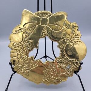 May include: A gold-toned metal Christmas wreath ornament with a bow and holly leaves. The wreath is on a black metal stand.