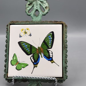 Puede incluir: Un soporte para posavasos de metal verde en forma de mariposa con una baldosa de cerámica blanca con tres mariposas. Las mariposas son verdes, azules y amarillas con marcas negras.