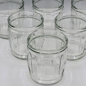 Könnte beinhalten: Sechs klare Glasbecher mit einem strukturierten, gerippten Design. Die Becher sind leer und in einem Halbkreis auf einer weißen Oberfläche angeordnet.