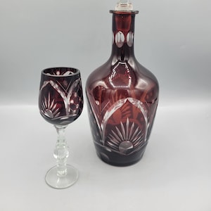 Puede incluir: Un juego de dos decantadores y copas de vino de vidrio vintage. El decantador es de color rojo oscuro con un tapón de vidrio transparente y diseños intrincados de vidrio cortado. La copa de vino también es de color rojo oscuro con diseños de vidrio cortado.