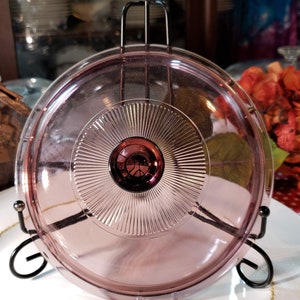 May include: A vintage pink glass lid with a raised, circular design and a dark pink knob in the center. The lid is on a black metal stand.