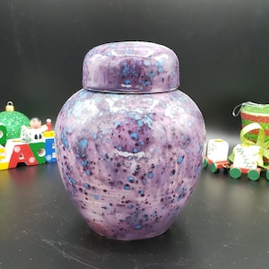 Könnte beinhalten: Eine violette Keramikdose mit einem weißen Deckel und blauen und weißen Flecken. Die Dose hat einen runden Körper und eine breite Öffnung.
