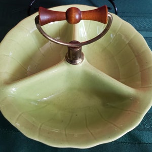 Puede incluir: Un plato de servir de tres secciones con un asa de madera. El plato es de color verde claro y tiene un borde festoneado. El asa está unida a una base de metal.