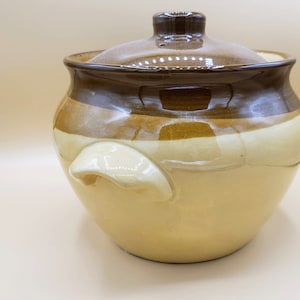 Puede incluir: Una olla de cerámica marrón y beige con tapa. La olla tiene un cuerpo redondeado y una sola asa en el lateral. La tapa tiene un pequeño pomo en la parte superior.