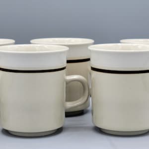 May include: Six beige ceramic mugs with a white rim and a brown stripe around the middle.