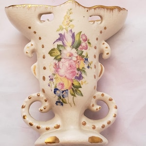 May include: A white ceramic vase with gold accents and a floral design. The vase has two handles and a scalloped top.