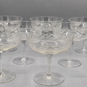 May include: Set of ten clear glass champagne coupes with a diamond pattern. The coupes are arranged in a semi-circle on a white surface.
