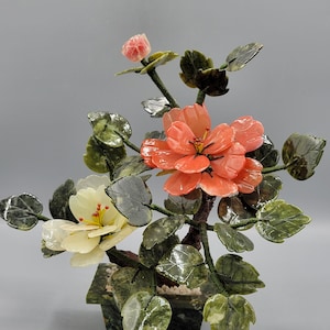 Op de afbeelding: Een jade bloemenarrangement met drie roze en witte bloemen en groene bladeren in een groene jade pot. De bloemen zijn gesneden uit jade en de bladeren zijn van jade.