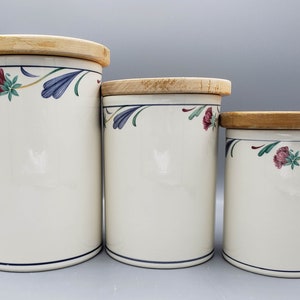 Könnte beinhalten: Set aus drei Küchenbehältern aus Keramik mit Holzdeckeln. Die cremefarbenen Behälter haben ein Blumenmuster in Rot, Blau und Grün. Jeder Behälter hat eine dunkelblaue Linie am Boden und einen Holzdeckel.