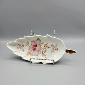 Puede incluir: Un plato de porcelana blanca con forma de hoja con un diseño floral rosa y verde. El plato tiene un borde dorado.