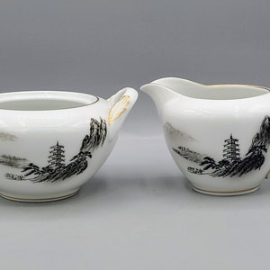 Może przedstawiać: Biały porcelanowy zestaw cukiernicy i mlecznika z czarnymi i złotymi malowanymi wzorami azjatyckich krajobrazów.