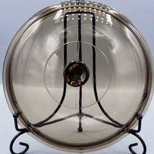 May include: A clear glass lid with a brown tint, resting on a black metal stand. The lid has a small knob on top.