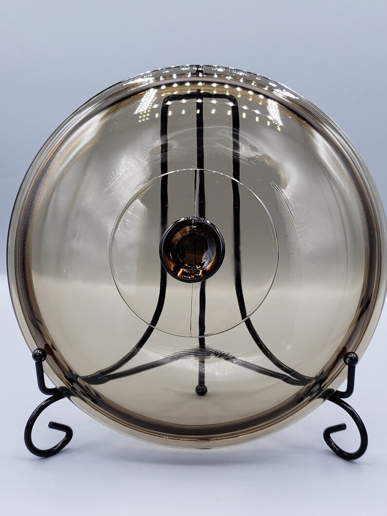 May include: A clear glass lid with a brown tint, resting on a black metal stand. The lid has a small knob on top.