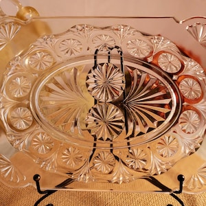 May include: A clear glass serving tray with a decorative, cut-glass design. The tray has an oval center and a square outer edge.