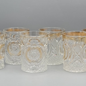 May include: Six clear glass tumblers with a gold rim and intricate etched designs. The glasses are arranged in a row on a grey surface.