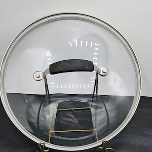 May include: A clear glass lid with a black handle and a silver rim. The lid is sitting on a gold metal stand with three legs.