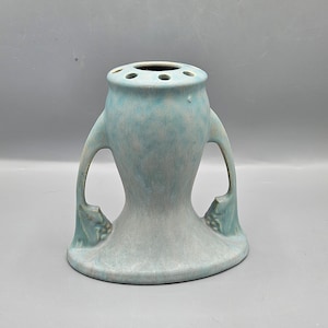 Könnte beinhalten: Eine hellblaue Keramikvase mit zwei Griffen und einem runden Boden. Die Vase hat eine strukturierte Oberfläche und einen Ring von Löchern um die Oberseite.