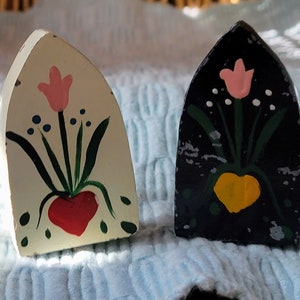Puede incluir: Dos pequeñas figuritas de madera pintadas a mano con forma de ventanas arqueadas. Una es blanca con un corazón rojo y una flor rosa, la otra es negra con un corazón amarillo y una flor rosa.
