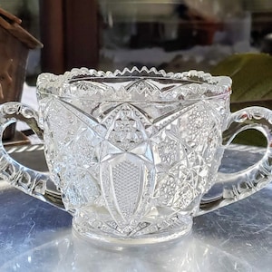 Può includere: Una zuccheriera di vetro trasparente con due manici. La ciotola ha un design in vetro tagliato ed è appoggiata su un vassoio d'argento.