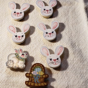 May include: Six wooden bunny rabbit figurines with painted faces and a wooden basket with painted Easter eggs. The bunnies are white with pink ears and noses. The basket is brown with blue, green, and pink eggs.