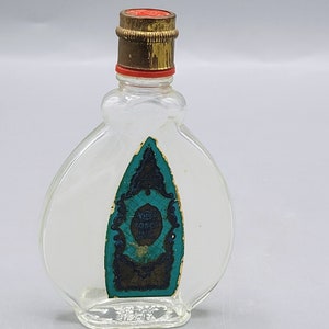Pode incluir: Um pequeno frasco de vidro transparente com uma tampa dourada e vermelha. O frasco tem um rótulo azul esverdeado e dourado com o texto "Tosca Eau de Cologne".