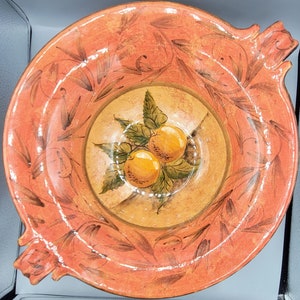 Puede incluir: Un cuenco de terracota con un diseño pintado a mano de naranjas y hojas. El cuenco tiene dos asas y un esmalte de color melocotón.